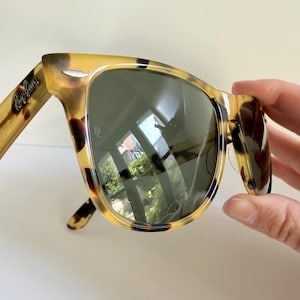 May include: Tortoiseshell-patterned sunglasses with dark lenses. The sunglasses have a classic design with the Ray-Ban logo visible on the temple. The lenses reflect the surrounding environment.