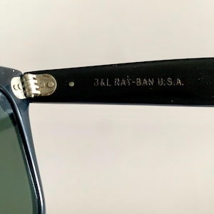 May include: Close-up of a pair of sunglasses with a dark navy frame and green lenses. The text "B&L RAY-BAN U.S.A." is printed on the black arm of the sunglasses. The hinge is silver.