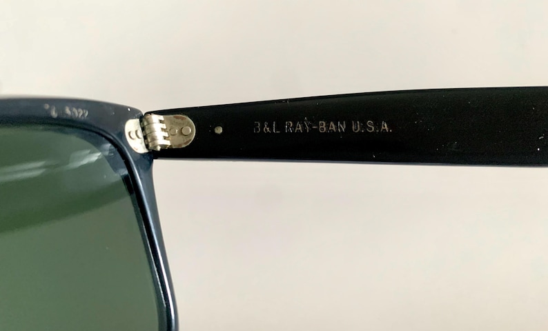 May include: Close-up of a pair of sunglasses with a dark navy frame and green lenses. The text "B&L RAY-BAN U.S.A." is printed on the black arm of the sunglasses. The hinge is silver.