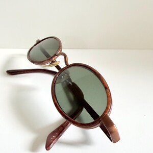 May include: Brown-framed sunglasses with oval lenses. The sunglasses have a classic design with a bridge and temples. The lenses are a dark green colour, and the frame has a glossy finish.