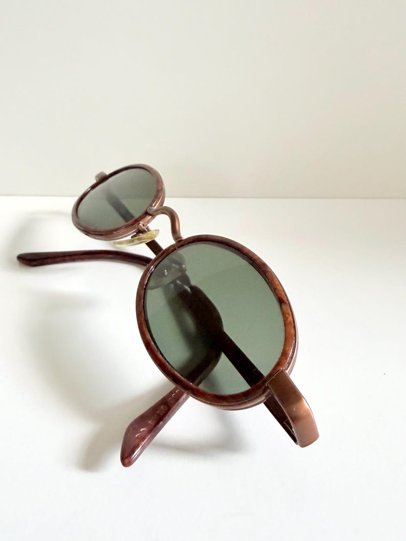 May include: Brown-framed sunglasses with oval lenses. The sunglasses have a classic design with a bridge and temples. The lenses are a dark green colour, and the frame has a glossy finish.