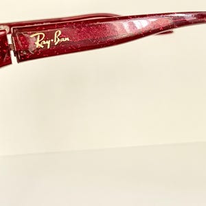 May include: Close-up of a pair of red-tinted sunglasses with the Ray-Ban logo in gold. The sunglasses have a glittery finish and a curved design. The lenses are reflective.
