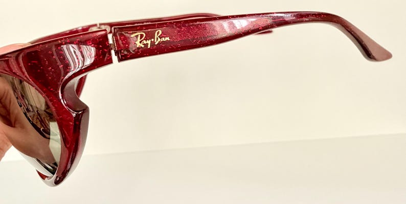 May include: Close-up of a pair of red-tinted sunglasses with the Ray-Ban logo in gold. The sunglasses have a glittery finish and a curved design. The lenses are reflective.