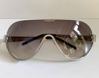 Puede incluir: Gafas de sol estilo máscara con lentes degradadas en marrón y montura plateada. El puente y el borde inferior están adornados con pequeños cristales transparentes. La palabra FERRE está grabada en la parte superior y en las bisagras.