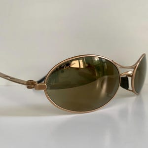 May include: Gold-tone Ray-Ban sunglasses with oval lenses and a sleek, modern design. The lenses have a reflective, mirrored finish. The Ray-Ban logo is visible on the lens. The temples are thin and gold, with black ear pieces.