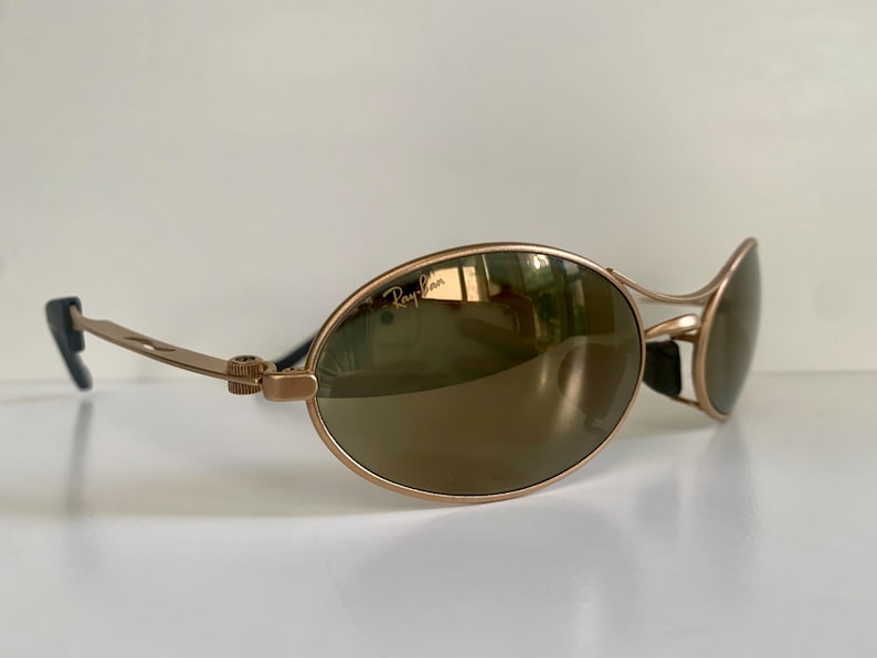 May include: Gold-tone Ray-Ban sunglasses with oval lenses and a sleek, modern design. The lenses have a reflective, mirrored finish. The Ray-Ban logo is visible on the lens. The temples are thin and gold, with black ear pieces.