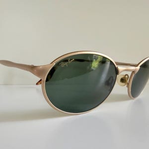 Puede incluir: Gafas de sol Ray-Ban doradas con lentes verde oscuro. Las monturas ovaladas tienen un brillo met&aacute;lico sutil, y las patillas presentan un patr&oacute;n de carey en las puntas. El logotipo de Ray-Ban es visible en la lente superior izquierda.