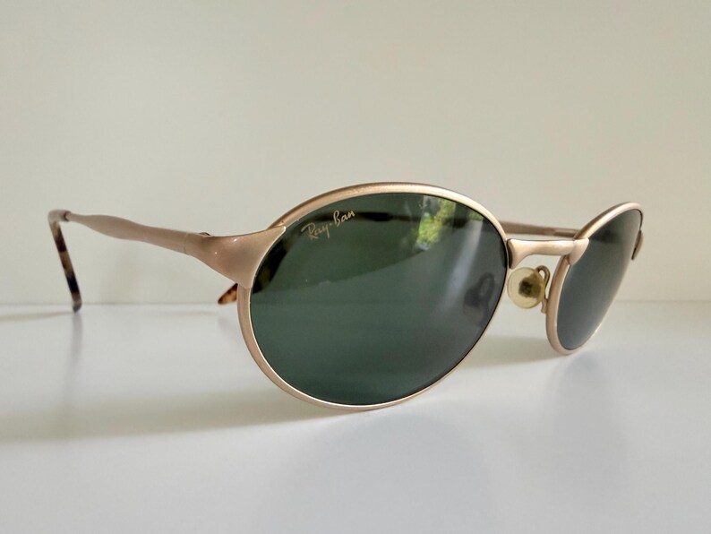 Puede incluir: Gafas de sol Ray-Ban doradas con lentes verde oscuro. Las monturas ovaladas tienen un brillo met&aacute;lico sutil, y las patillas presentan un patr&oacute;n de carey en las puntas. El logotipo de Ray-Ban es visible en la lente superior izquierda.