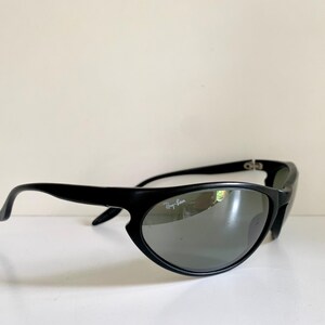 May include: Black sunglasses with a wrap-around frame design and dark, reflective lenses. The right lens features the white Ray-Ban logo printed in the upper corner. The frames have a smooth, matte finish with curved temples and metal hinges.