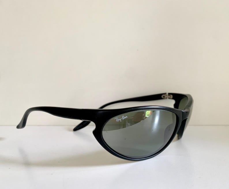May include: Black sunglasses with a wrap-around frame design and dark, reflective lenses. The right lens features the white Ray-Ban logo printed in the upper corner. The frames have a smooth, matte finish with curved temples and metal hinges.