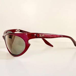 May include: A pair of Ray-Ban sunglasses with a unique, curved design. The frames are a sparkling burgundy color, and the lenses are a reflective gray. The Ray-Ban logo is visible on the temple.