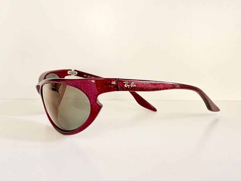 May include: A pair of Ray-Ban sunglasses with a unique, curved design. The frames are a sparkling burgundy color, and the lenses are a reflective gray. The Ray-Ban logo is visible on the temple.