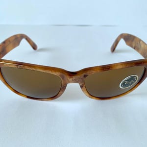 Puede incluir: Gafas de sol Ray-Ban de carey marr&oacute;n con lentes marrones. Las gafas tienen una barra de metal sobre el puente de la nariz y un logotipo de Ray-Ban en la lente derecha.