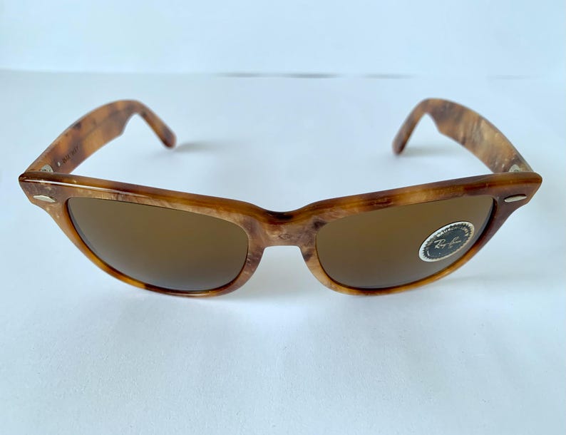 Puede incluir: Gafas de sol Ray-Ban de carey marr&oacute;n con lentes marrones. Las gafas tienen una barra de metal sobre el puente de la nariz y un logotipo de Ray-Ban en la lente derecha.