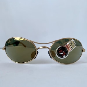 May include: Gold-framed sunglasses with oval lenses and a bridge. The lenses are a muted green color. A sticker with text and Olympic rings is affixed to the right lens. The Ray-Ban logo is visible on the left lens.