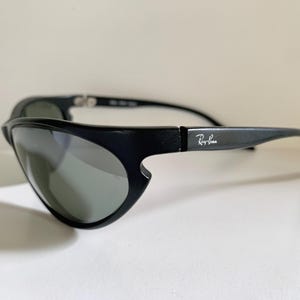 May include: Black Ray-Ban sunglasses featuring a wrap-around frame design with dark tinted lenses. The temples display the white Ray-Ban logo on the side. The frame has a distinct cutout detail near the lens edge and a smooth matte finish.