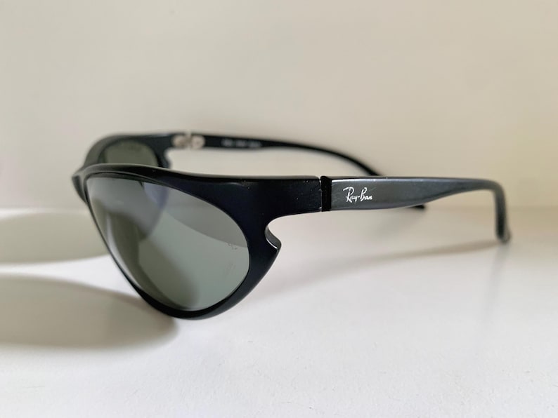 May include: Black Ray-Ban sunglasses featuring a wrap-around frame design with dark tinted lenses. The temples display the white Ray-Ban logo on the side. The frame has a distinct cutout detail near the lens edge and a smooth matte finish.