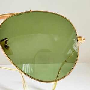 May include: Close-up of gold-rimmed sunglasses with green lenses. The sunglasses have a classic aviator design, featuring a double bridge and curved temples. The lenses are a light green color, and the frame is a shiny gold.