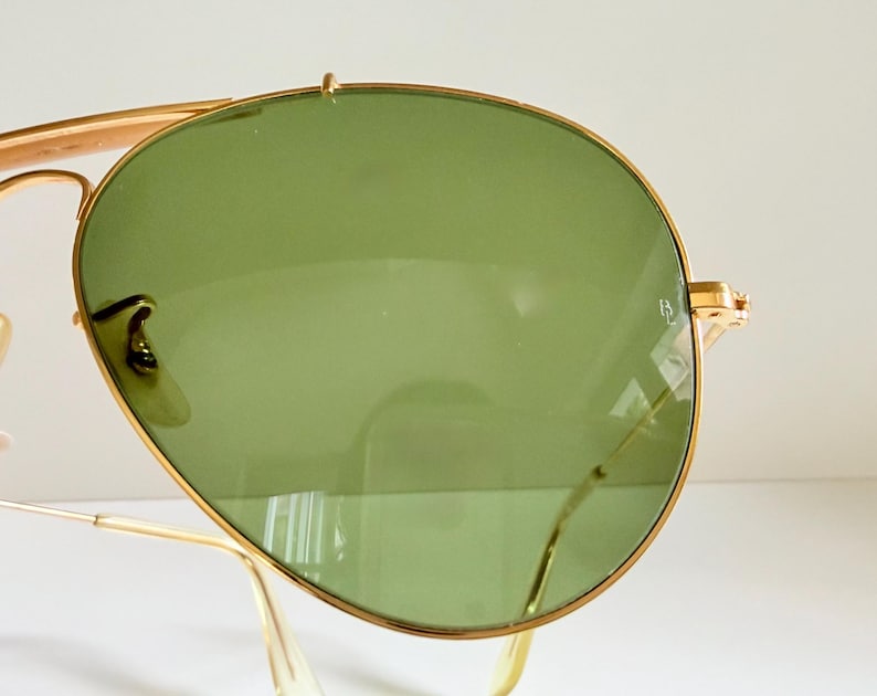 May include: Close-up of gold-rimmed sunglasses with green lenses. The sunglasses have a classic aviator design, featuring a double bridge and curved temples. The lenses are a light green color, and the frame is a shiny gold.