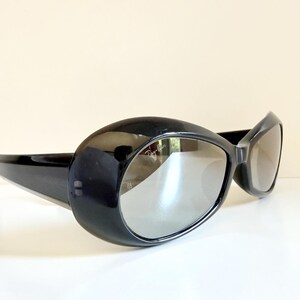 May include: Black oversized sunglasses with a glossy finish. The sunglasses have a rounded, oval shape with thick frames and reflective lenses. The arms are sleek and extend back to the temples.