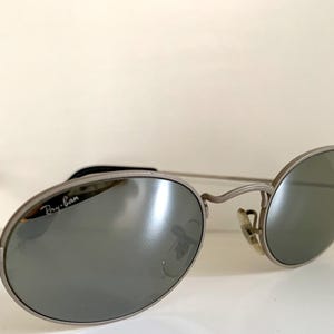 Puede incluir: Gafas de sol Ray-Ban plateadas con lentes de forma ovalada y acabado espejado. Las gafas tienen un fino marco de metal y el logotipo de Ray-Ban es visible en la lente derecha.