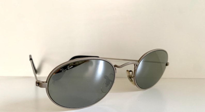 Puede incluir: Gafas de sol Ray-Ban plateadas con lentes de forma ovalada y acabado espejado. Las gafas tienen un fino marco de metal y el logotipo de Ray-Ban es visible en la lente derecha.