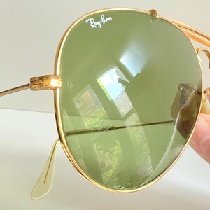 May include: Close-up of gold-rimmed Ray-Ban sunglasses with green lenses. The Ray-Ban logo is visible on the top left lens. The sunglasses have a classic aviator style with a double bridge and thin temples.