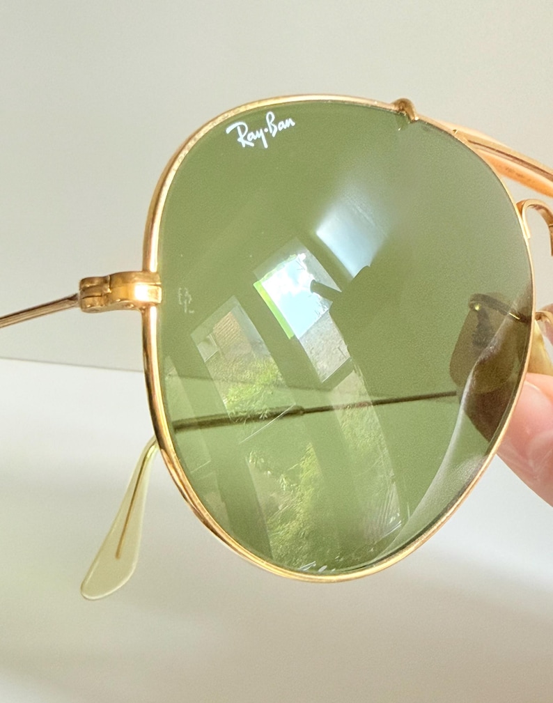May include: Close-up of gold-rimmed Ray-Ban sunglasses with green lenses. The Ray-Ban logo is visible on the top left lens. The sunglasses have a classic aviator style with a double bridge and thin temples.