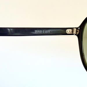 May include: Black sunglasses with green-tinted lenses. The frame is black with the word "SHELBY" printed on the temple. The sunglasses have a classic, rounded shape.