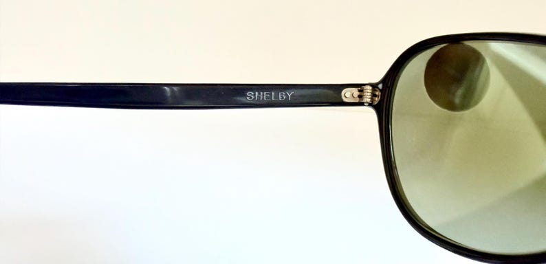 May include: Black sunglasses with green-tinted lenses. The frame is black with the word "SHELBY" printed on the temple. The sunglasses have a classic, rounded shape.