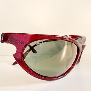 May include: A pair of vintage Ray-Ban sunglasses with a unique, angular design. The frames are a translucent, speckled burgundy color, and the lenses are a muted green. The Ray-Ban logo is visible on the lens.