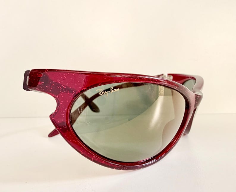 May include: A pair of vintage Ray-Ban sunglasses with a unique, angular design. The frames are a translucent, speckled burgundy color, and the lenses are a muted green. The Ray-Ban logo is visible on the lens.
