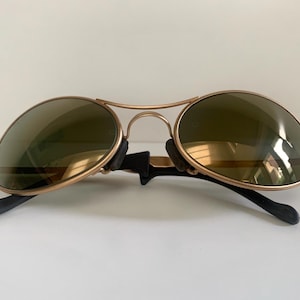 May include: Gold-framed aviator sunglasses with olive-green lenses. The sunglasses have a double bridge and black earpieces. The lenses are oval-shaped and reflect light. A classic eyewear accessory.