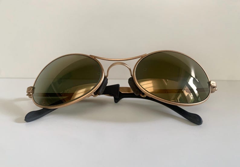 May include: Gold-framed aviator sunglasses with olive-green lenses. The sunglasses have a double bridge and black earpieces. The lenses are oval-shaped and reflect light. A classic eyewear accessory.