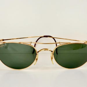 K&ouml;nnte beinhalten: Goldfarbene Sonnenbrille mit gr&uuml;nen Gl&auml;sern. Die Brille hat einen Doppelsteg und einen Brauensteg. Die Gl&auml;ser sind oval geformt. Der Markenname ist auf dem linken Glas sichtbar.