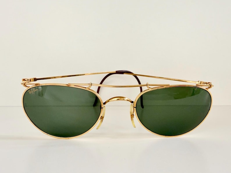 K&ouml;nnte beinhalten: Goldfarbene Sonnenbrille mit gr&uuml;nen Gl&auml;sern. Die Brille hat einen Doppelsteg und einen Brauensteg. Die Gl&auml;ser sind oval geformt. Der Markenname ist auf dem linken Glas sichtbar.