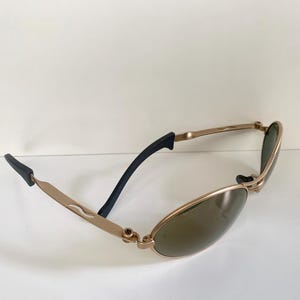 May include: Gold-toned sunglasses with oval lenses and black temple tips. The frames have a sleek, modern design. The lenses are a dark green color, providing shade and style.