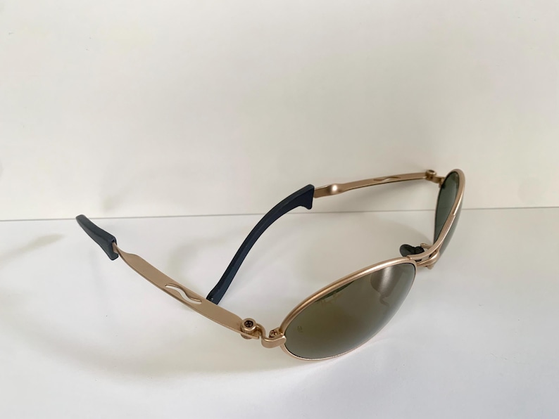 May include: Gold-toned sunglasses with oval lenses and black temple tips. The frames have a sleek, modern design. The lenses are a dark green color, providing shade and style.