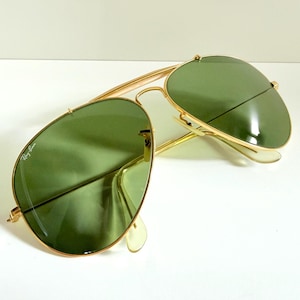 May include: Gold-framed aviator sunglasses with green lenses. The sunglasses have a classic teardrop shape and a double bridge. The Ray-Ban logo is visible on the left lens. The sunglasses are set against a white background.