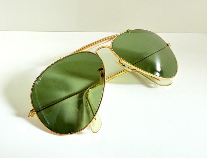 May include: Gold-framed aviator sunglasses with green lenses. The sunglasses have a classic teardrop shape and a double bridge. The Ray-Ban logo is visible on the left lens. The sunglasses are set against a white background.