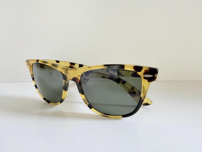 May include: Sunglasses with a classic design. The frames have a tortoise shell pattern in shades of yellow and brown. The lenses are a dark gray color. The sunglasses are set against a white background.