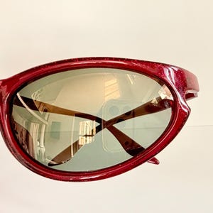 May include: Close-up of a pair of red sunglasses with a glittery finish. The lenses are a light green color, and the arms are brown. The sunglasses reflect the surrounding environment.