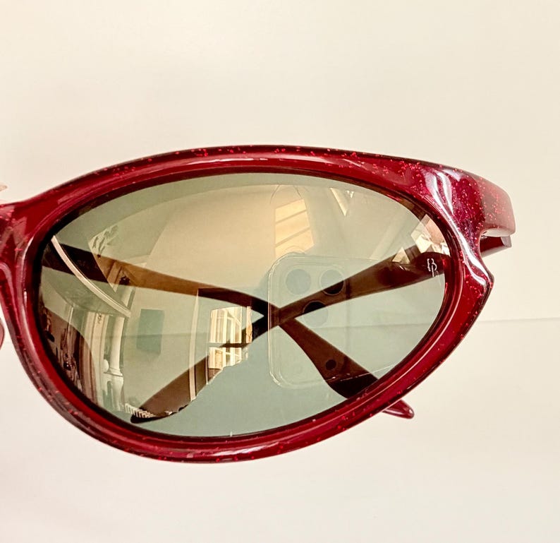 May include: Close-up of a pair of red sunglasses with a glittery finish. The lenses are a light green color, and the arms are brown. The sunglasses reflect the surrounding environment.