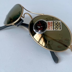 May include: Vintage gold-framed sunglasses with green lenses. The sunglasses have a classic aviator design with a double bridge and black temples. A sticker on the lens reads "SPORT 6-20 CM LENS" and features the Olympic rings.