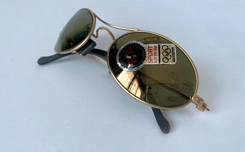 May include: Vintage gold-framed sunglasses with green lenses. The sunglasses have a classic aviator design with a double bridge and black temples. A sticker on the lens reads "SPORT 6-20 CM LENS" and features the Olympic rings.