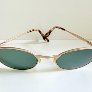 Puede incluir: Gafas de sol doradas vintage con lentes ovaladas verdes. Las monturas tienen un dise&ntilde;o delgado y elegante con puente y varillas. Las varillas presentan un patr&oacute;n de carey. El texto "B&L RAY-BAN" es visible.