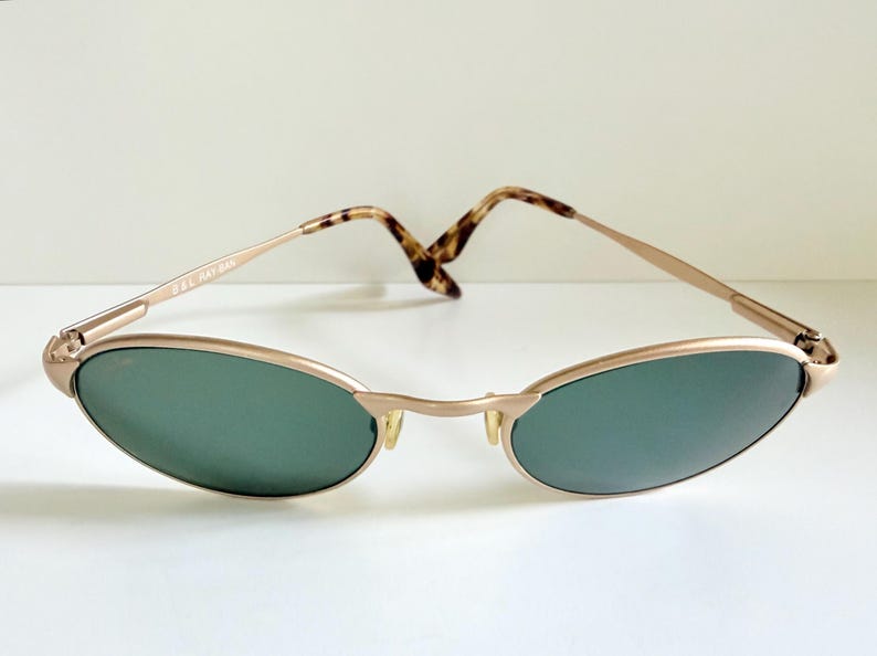 Puede incluir: Gafas de sol doradas vintage con lentes ovaladas verdes. Las monturas tienen un dise&ntilde;o delgado y elegante con puente y varillas. Las varillas presentan un patr&oacute;n de carey. El texto "B&L RAY-BAN" es visible.