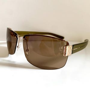 May include: Gucci sunglasses featuring rimless brown gradient lenses and olive green temples. The hinges are accented with a gold-tone metal piece displaying the repeating Gucci logo pattern. The brand name Gucci is engraved on the temple arm.