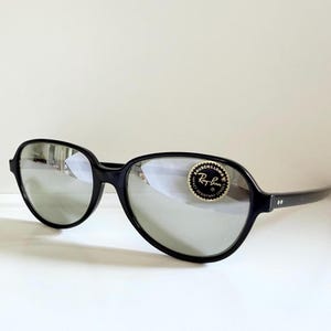 May include: Black Ray-Ban sunglasses with silver mirrored lenses. The sunglasses have a rounded, oversized frame and a small, circular sticker on the right lens. The arms of the sunglasses are thin and black.