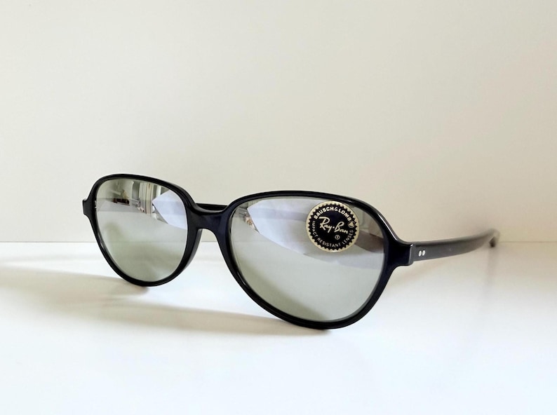 May include: Black Ray-Ban sunglasses with silver mirrored lenses. The sunglasses have a rounded, oversized frame and a small, circular sticker on the right lens. The arms of the sunglasses are thin and black.